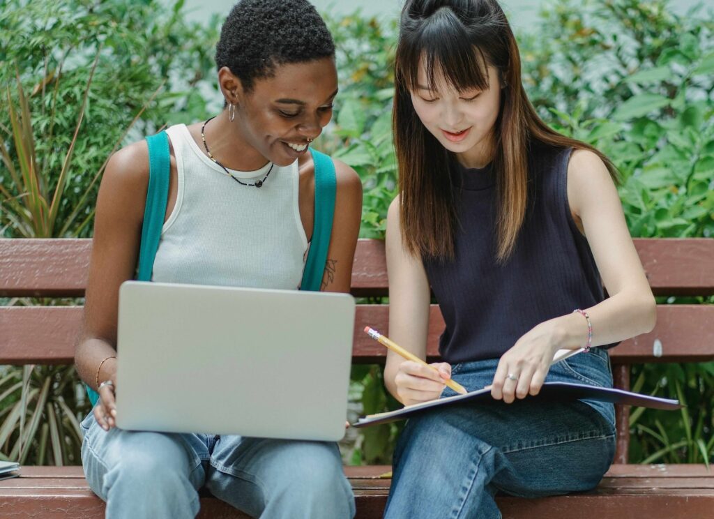 Two people sitting on a wooden bench outdoors, collaborating on a laptop and writing notes in a notebook, with green plants in the background.