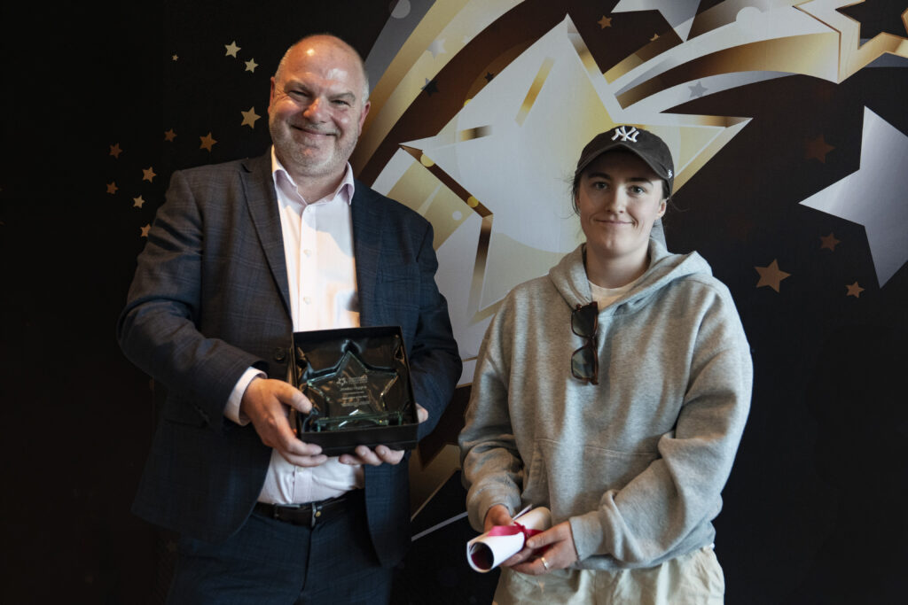 A smiling man in a suit holds a boxed award, standing next to a person in a grey hoodie and baseball cap holding a rolled-up certificate, against a star-patterned background.