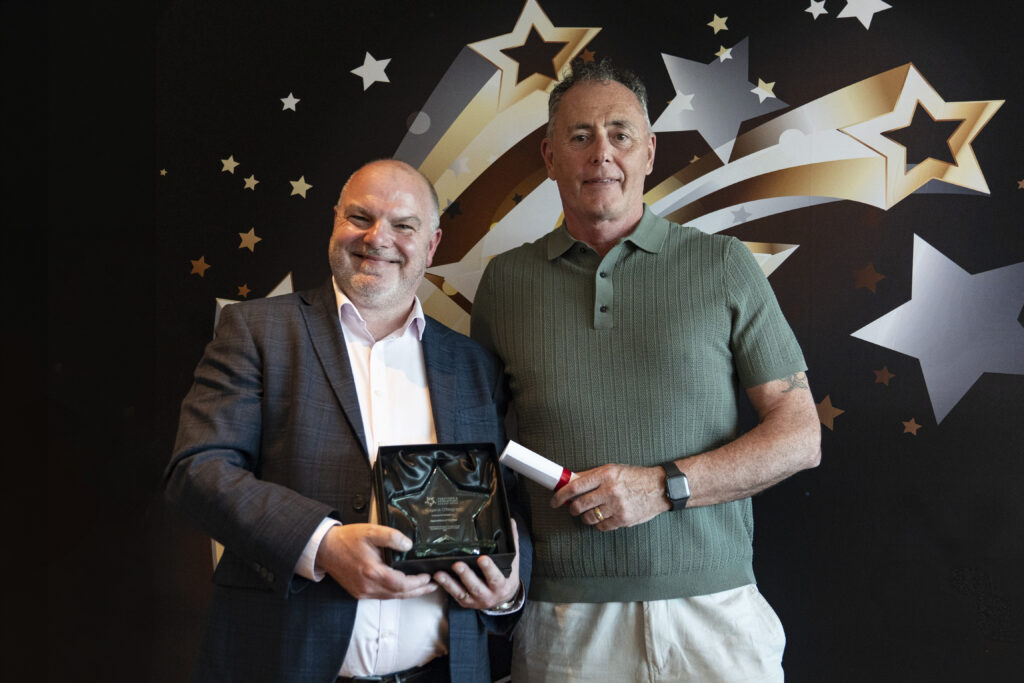 A smiling man in a suit holds a boxed award, standing next to a tall man green polo top holding a rolled-up certificate, against a star-patterned background.