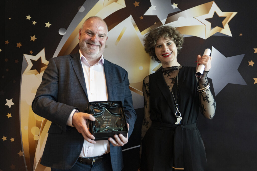 A smiling man in a suit holds a boxed award, standing next to a smiling woman with short curly hair in a black lace top and dress holding a rolled-up certificate, against a star-patterned background.