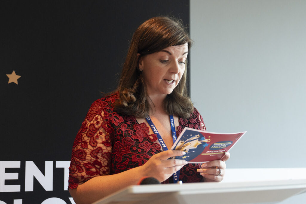 A woman with long dark hair and a red lace top, wearing a "STAFF" lanyard, stands at a podium reading from an award booklet.