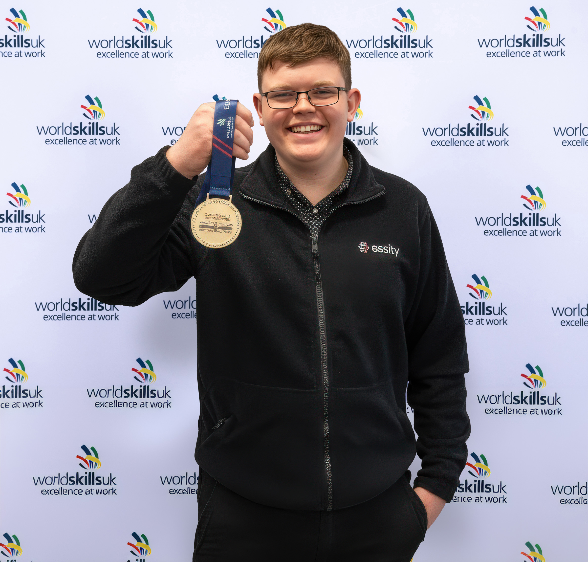 Edward standing against a world skills background with a bronze medal