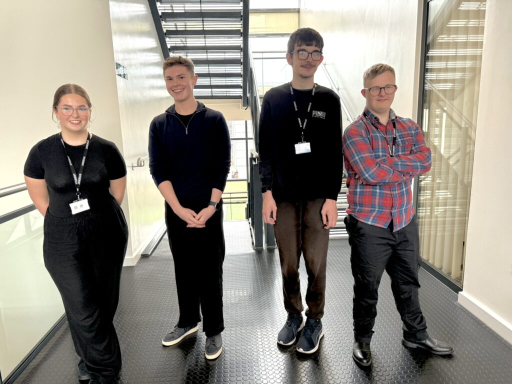 Four individuals wearing name badges standing side by side in a hallway with a staircase in the background.