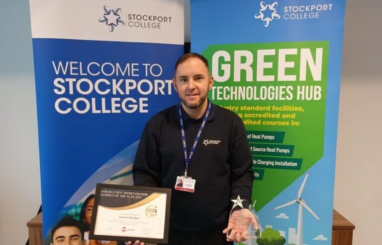 Jason Gallagher holding an award certificate and trophy at Stockport College, standing between banners for Stockport College and Green Technologies Hub.