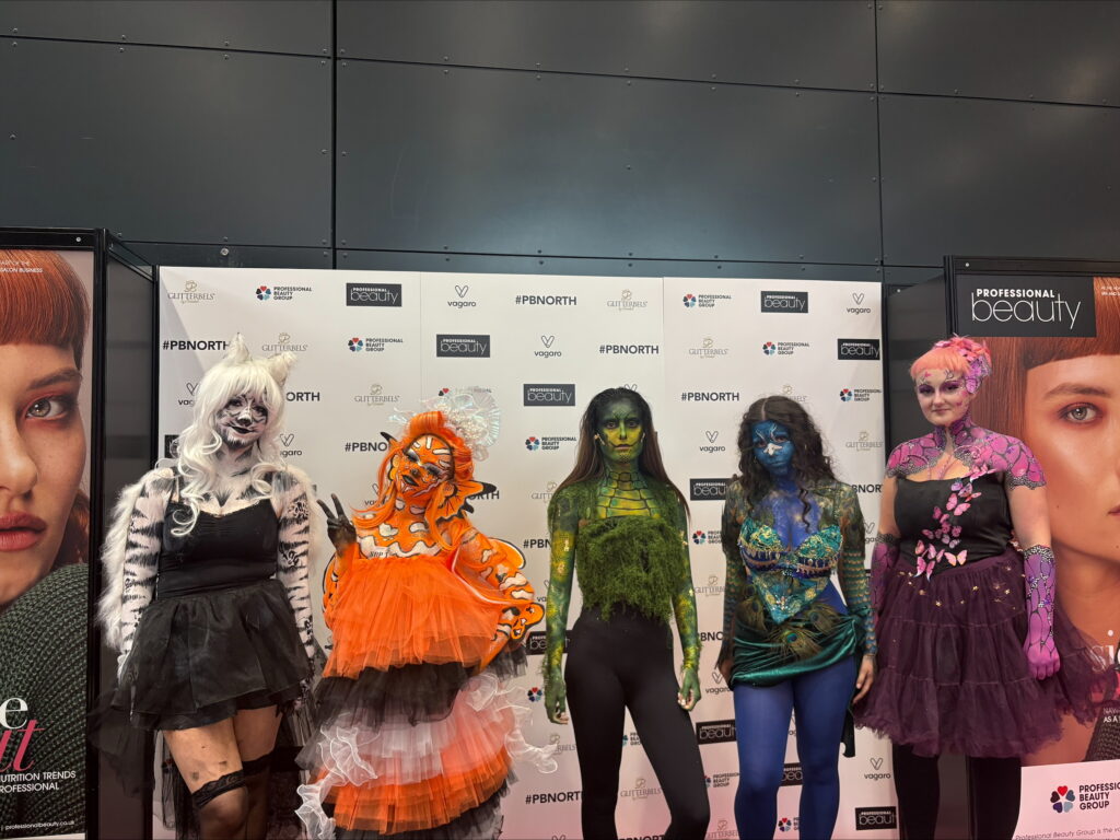 Five individuals in elaborate body paint and costumes posing in front of a branded backdrop at Professional Beauty North event