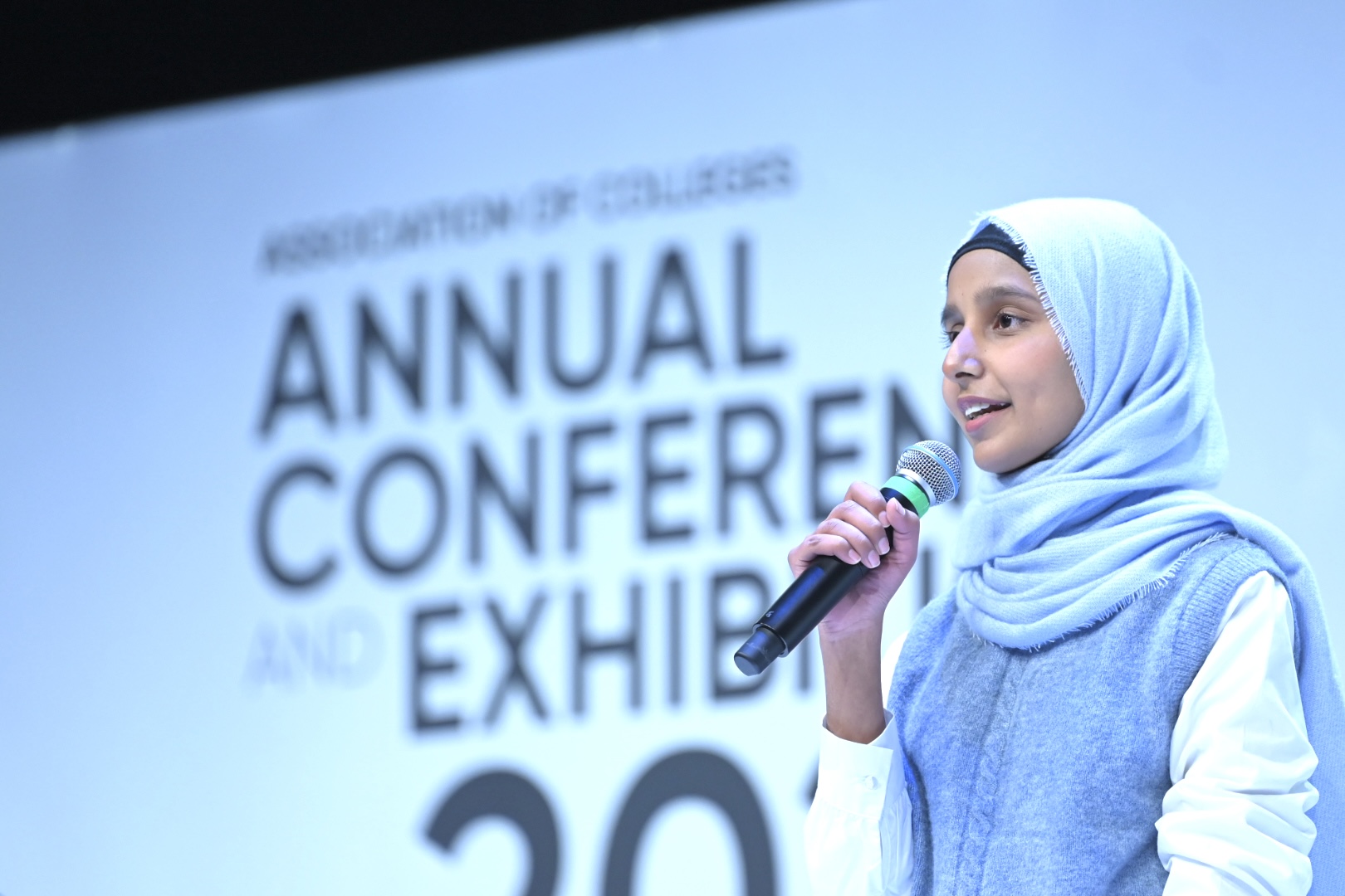 Hajira Qaiser speaking on stage at the Association of Colleges Annual Conference, holding a microphone in front of a large event backdrop