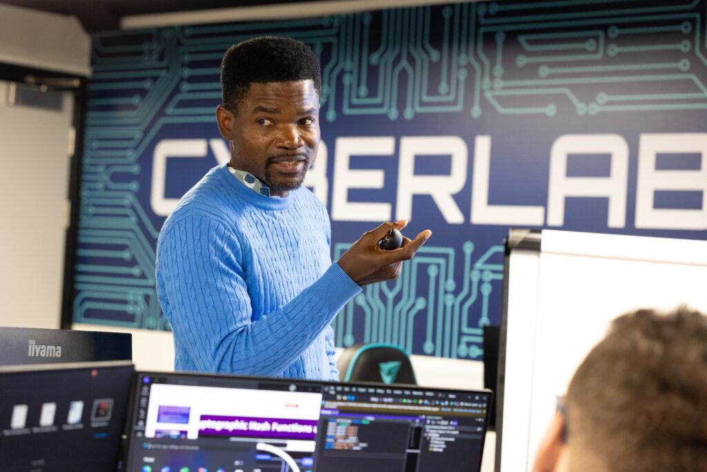 Instructor delivering a cybersecurity lecture in our brand new cyberlab, with coding applications visible on screens.