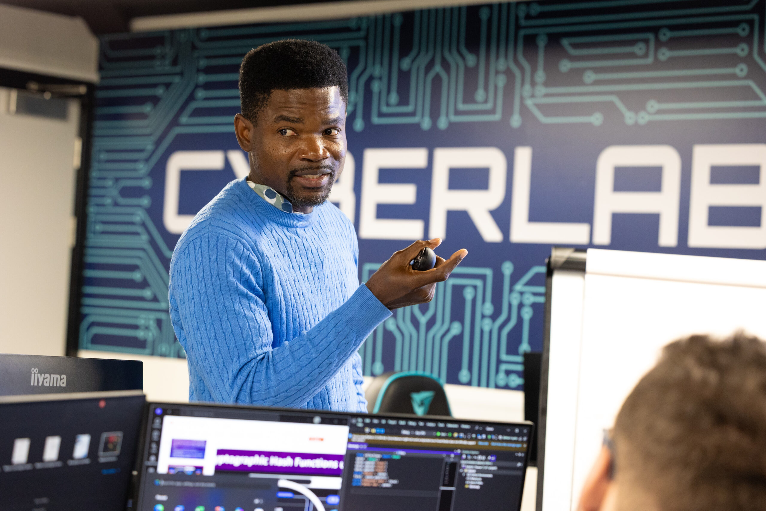 Instructor delivering a cybersecurity lecture in our brand new cyberlab, with coding applications visible on screens.