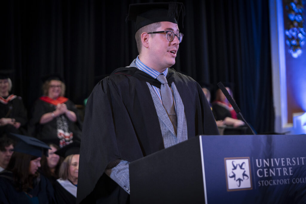 Graduate in academic regalia stands at the podium delivering a speech during the university graduation ceremony at University Centre Stockport College.