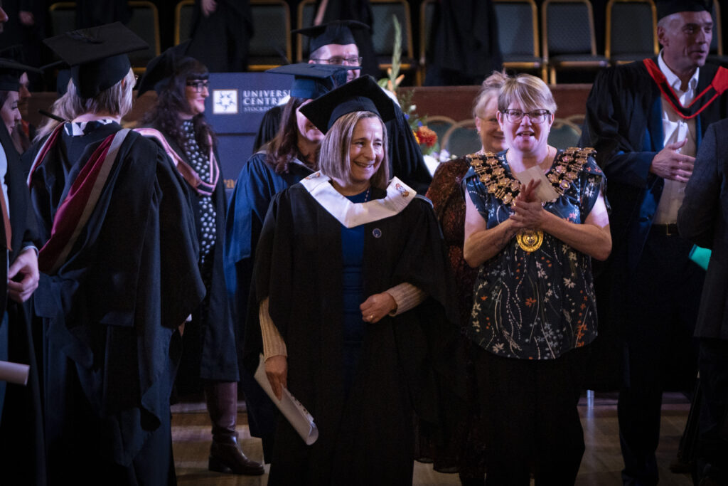 Academic staff in academic gowns with the Mayoress of Stockport wearing a ceremonial chain gather on stage during a university graduation ceremony.