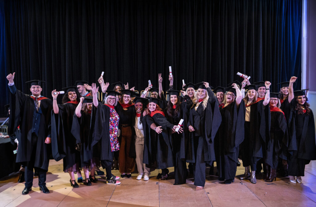 Group of graduates in academic gowns holding diplomas pose together on stage during the university graduation ceremony at University Centre Stockport College.