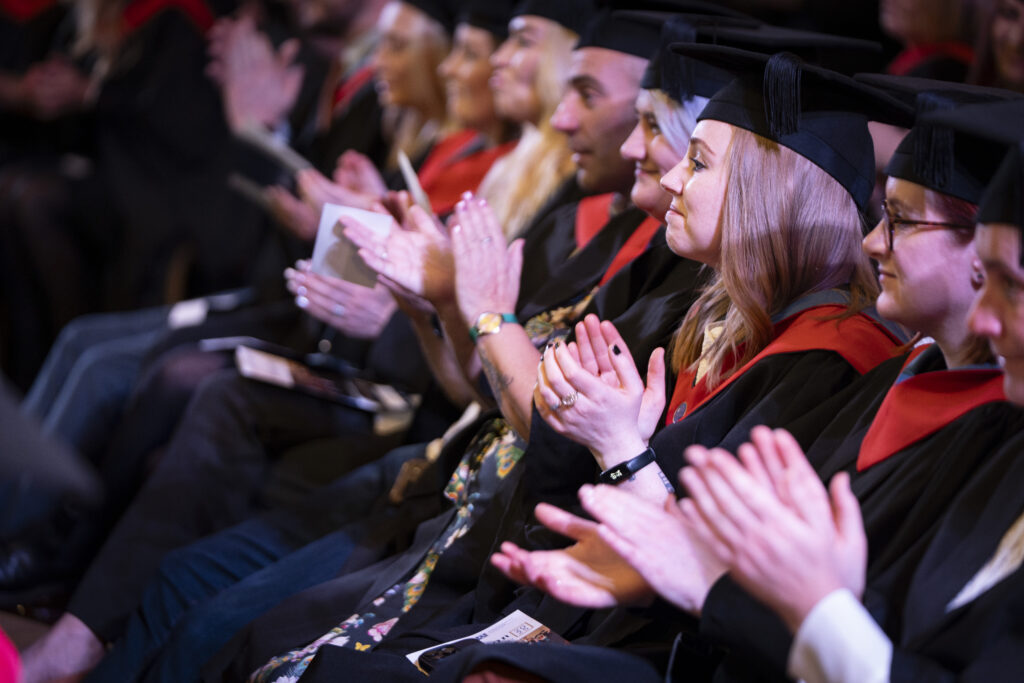 Graduates seated in rows wearing black gowns and mortarboards applaud during the university graduation ceremony at University Centre Stockport College.