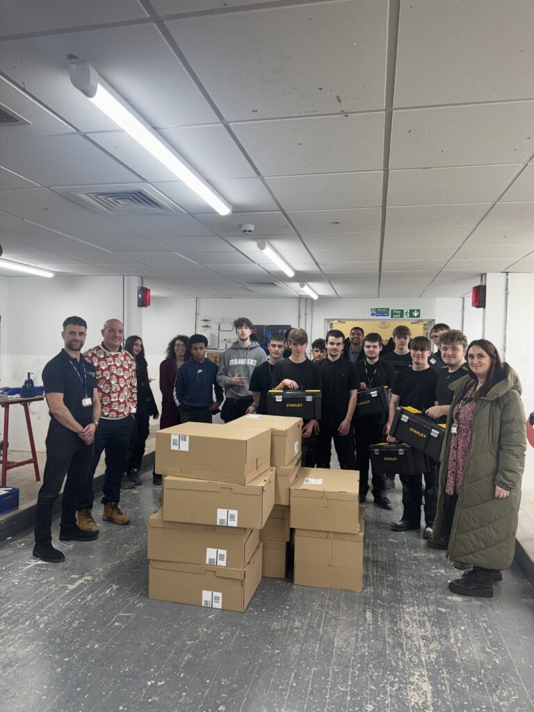 A group of people standing in a workshop. Several cardboard boxes are stacked in the foreground, and some students are holding black Stanley toolkits.
