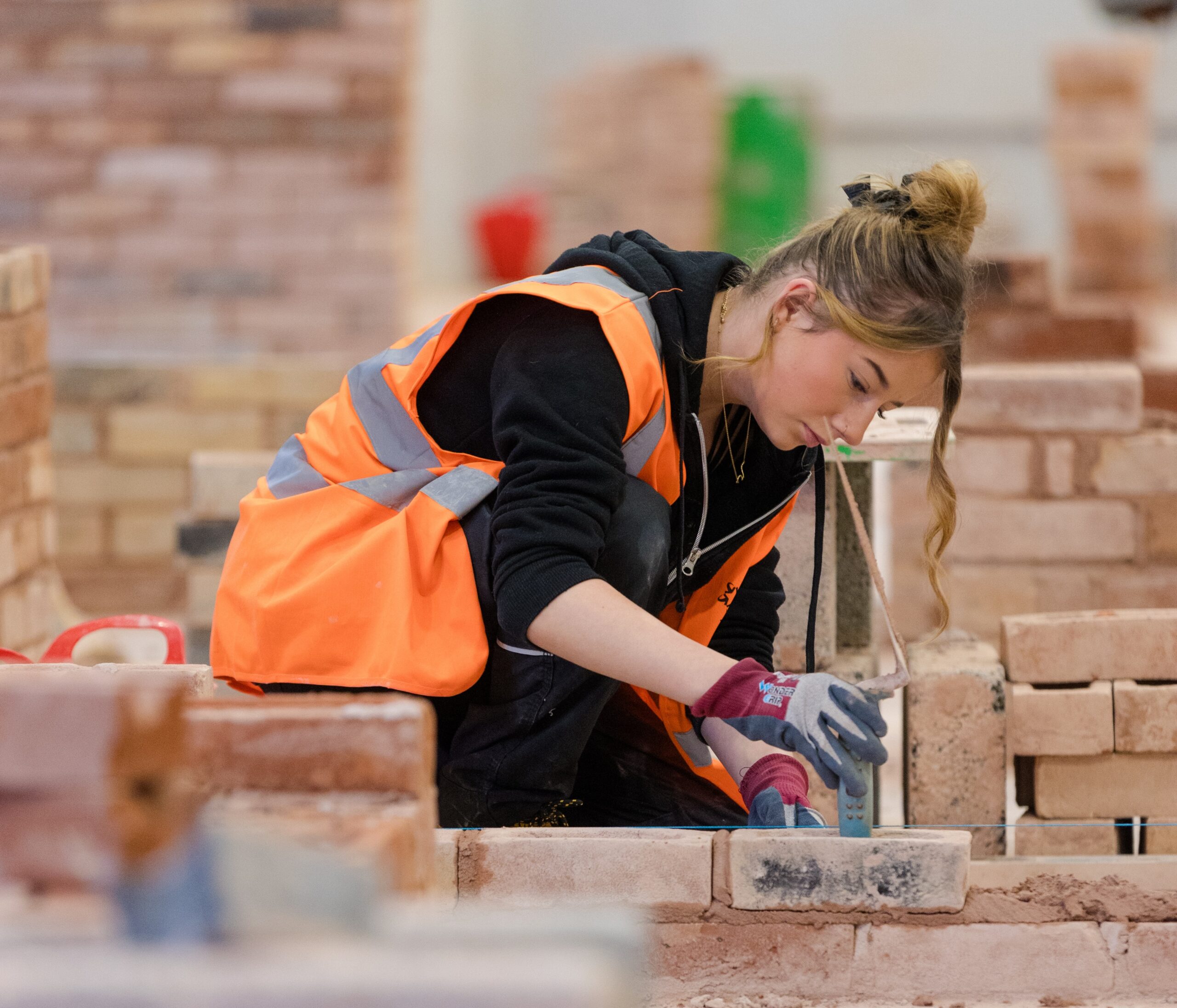 Young woman in an orange high vis jacket working on laying a brick wall, within a college workshop environment.