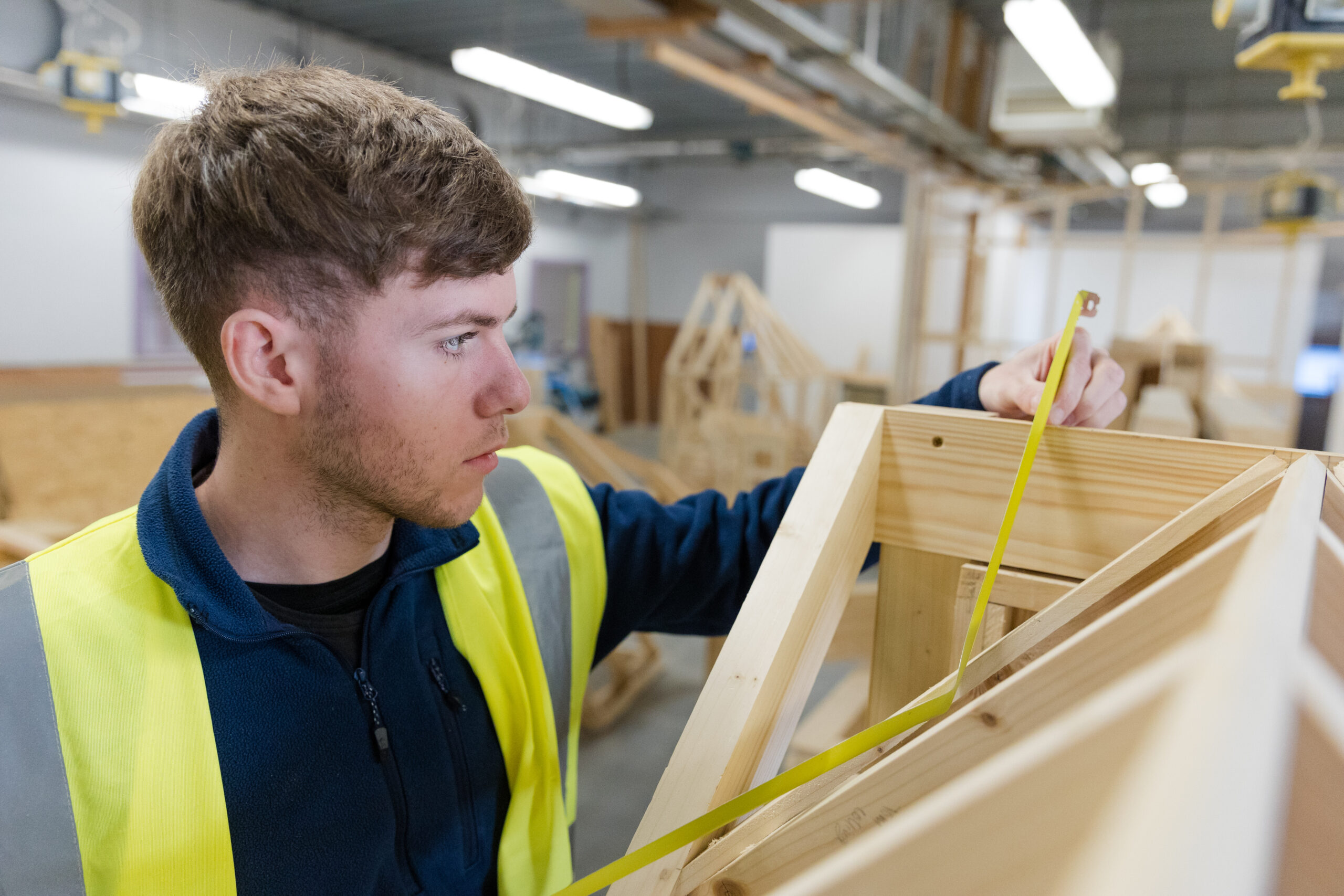 Young man wearing a his vis jacket in a workshop environment. He is focused on measuring a wooden structure using a yellow metal tape measure.