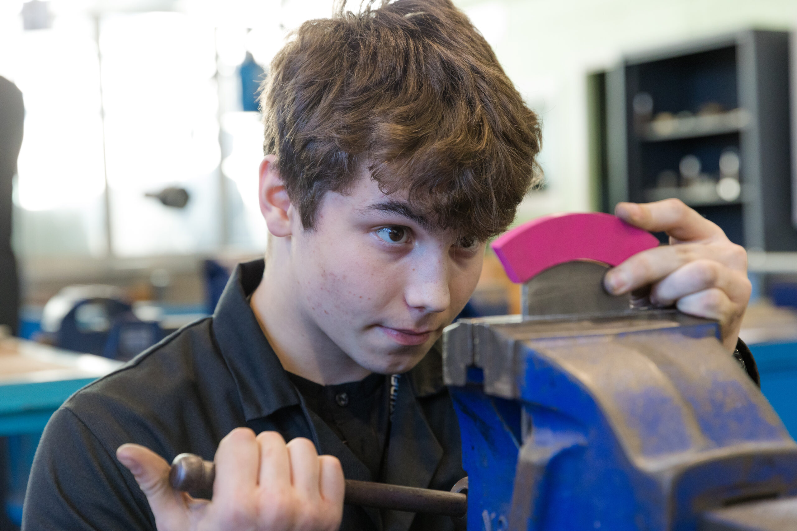 Young male wearing black work overalls in a workshop environment. He is deeply focused in a piece of metal equipment that he is looking at.