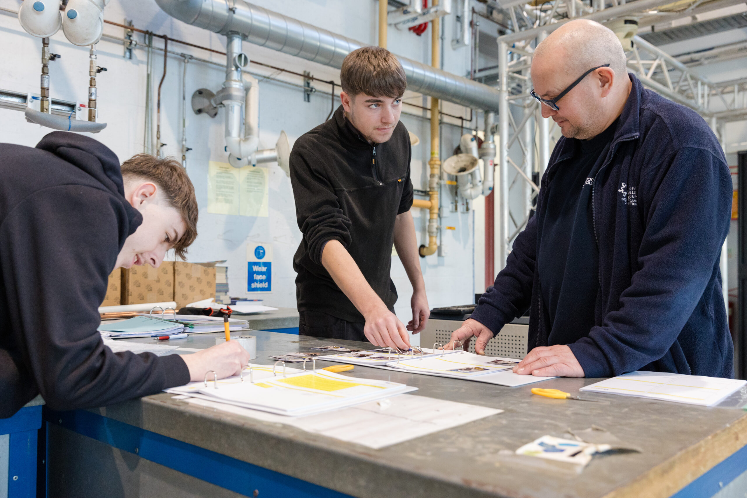 Three men gather around a table in a workshop environment. The two younger men are working on sheets within ringbinder folders, whilst the older man reviews the work in one of the folders.