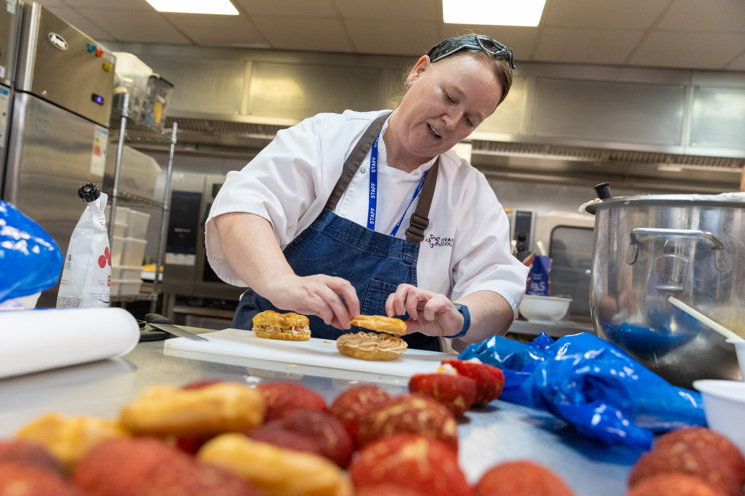 A adult woman dressed in chef clothing and a denim apron making a sandwich whilst stood in a commercial kitchen setting.