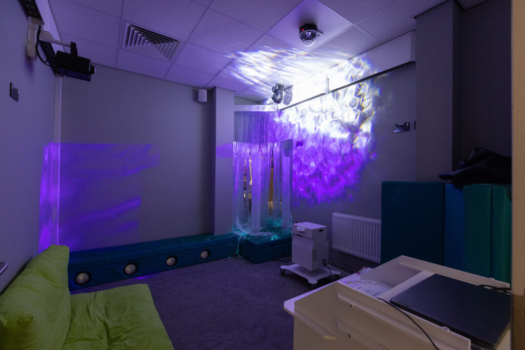 Sensory room featuring bubble tubes, colorful light projections on the walls and ceiling, cushioned seating, and a calming environment designed for relaxation and stimulation.