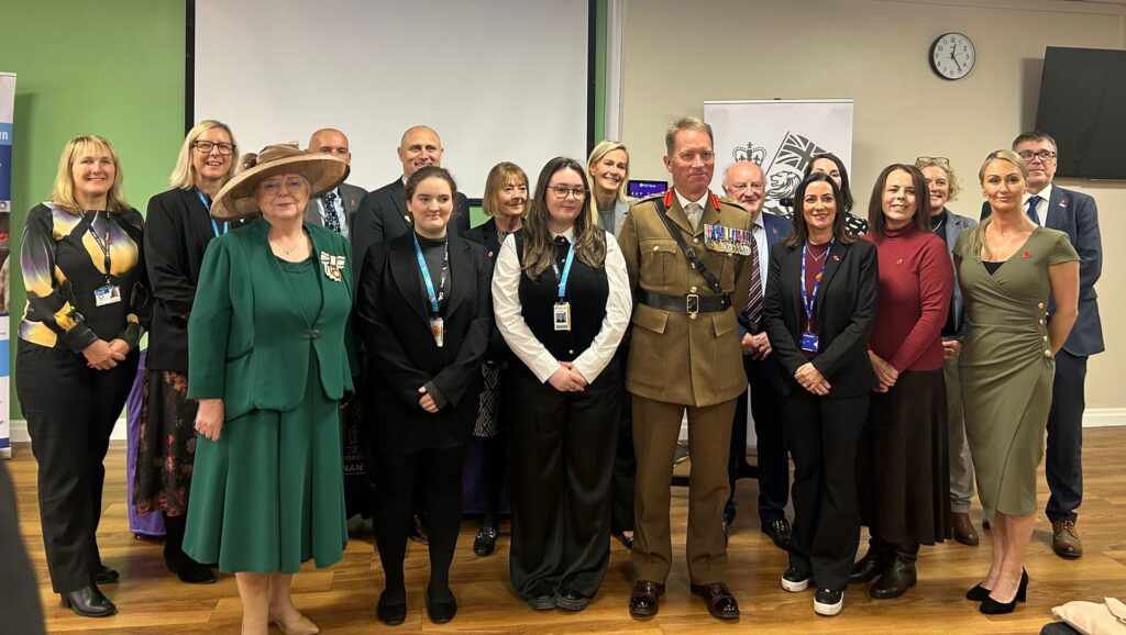 A group photo taken at a Covenant Signing Ceremony featuring a military representative and college staff standing in two rows. A presentation area with banners and screens is visible behind them.