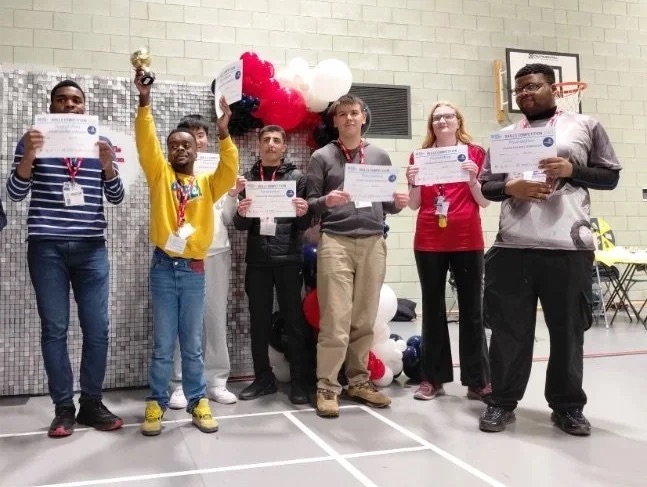 A group of learners celebrating at the GMC Skills Competition, holding certificates and a trophy in front of an event backdrop.